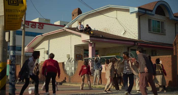 Zombies roam in a street while two men sitting on the roof of a school look at them