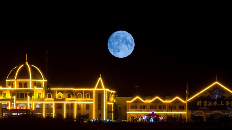  A bright full moon rises above a building lined with orange led lights. 