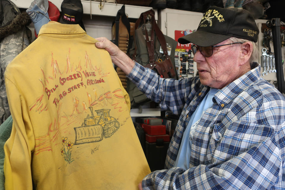 In this photograph taken Tuesday, April 26, 2022, outside Flagstaff, Ariz., Bill Velie displays a shirt that points to his career running a bulldozer to help contain wildfires. A massive wildfire that started Easter Sunday burned about 30 square miles and more than a dozen homes, hopscotching across the parched landscape. (AP Photo/Felicia Fonseca)