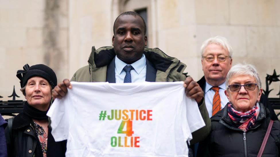 Oliver Campbell, dressed in a suit, stands outside the Royal Courts of Justice in London. He is surrounded on both sides by activists defending his case. He holds a white t-shirt with the slogan #justice4Ollie