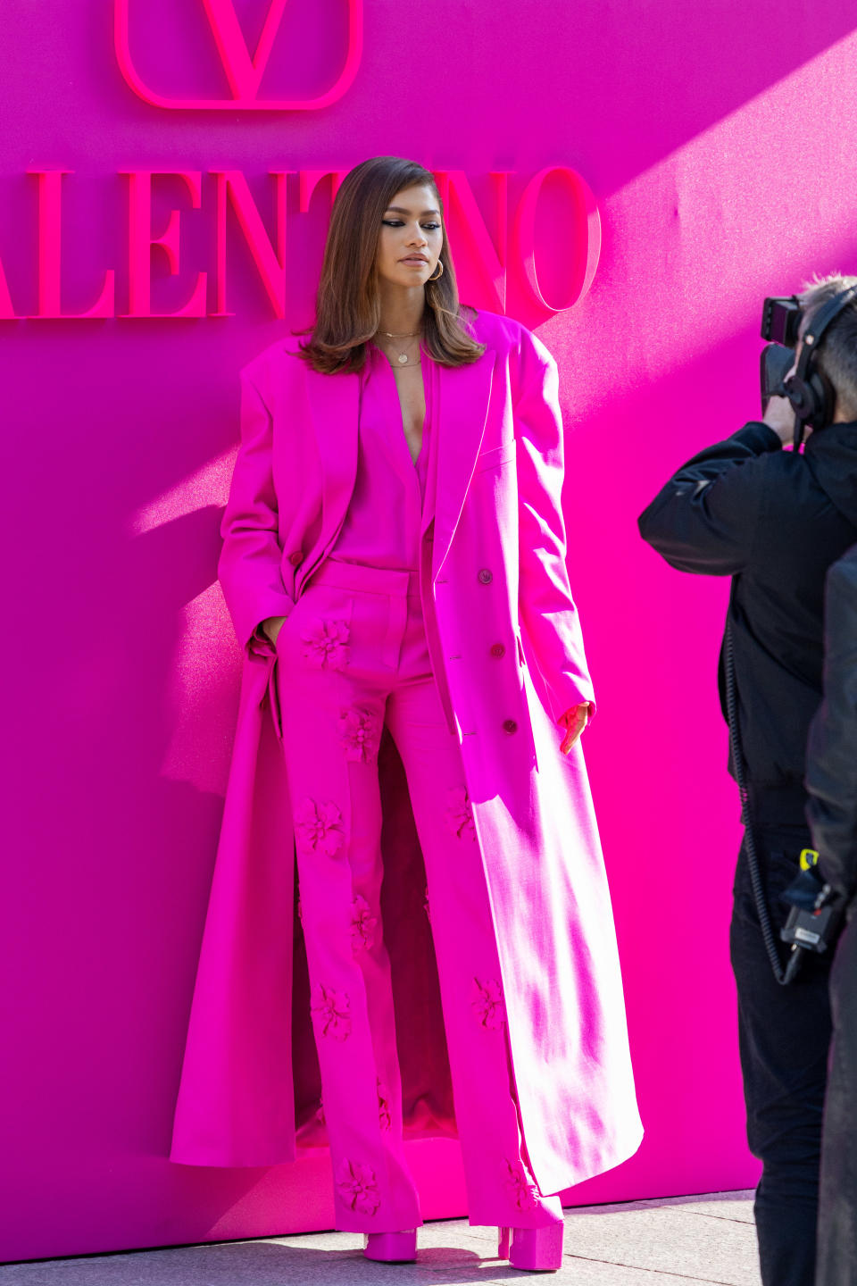 Zendaya attends the Valentino Womenswear Fall/Winter 2022/2023. She wears a bright fuchsia suit with matching tall platform heels and a matching ankle-length trenchcoat. The trousers have textured floral details.