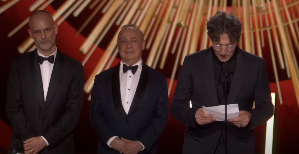 Three men in black tuxedos onstage, one reading from a paper with a microphone, flanked by the other two