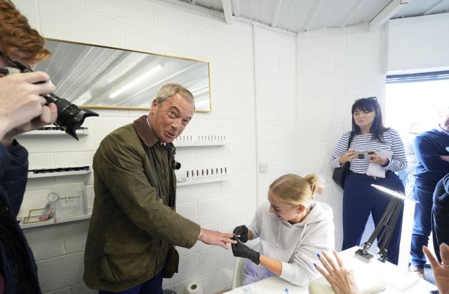 Nigel Farage holds his hand out in a nail salon while a woman in a grey hoodie and black rubber glove paints his finger while a camera takes his picture