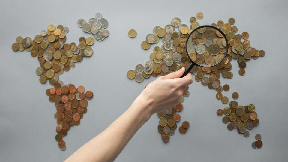 Eine Person hält eine Lupe über ein mit Münzen geschaffenes Abbild der Weltkarte
