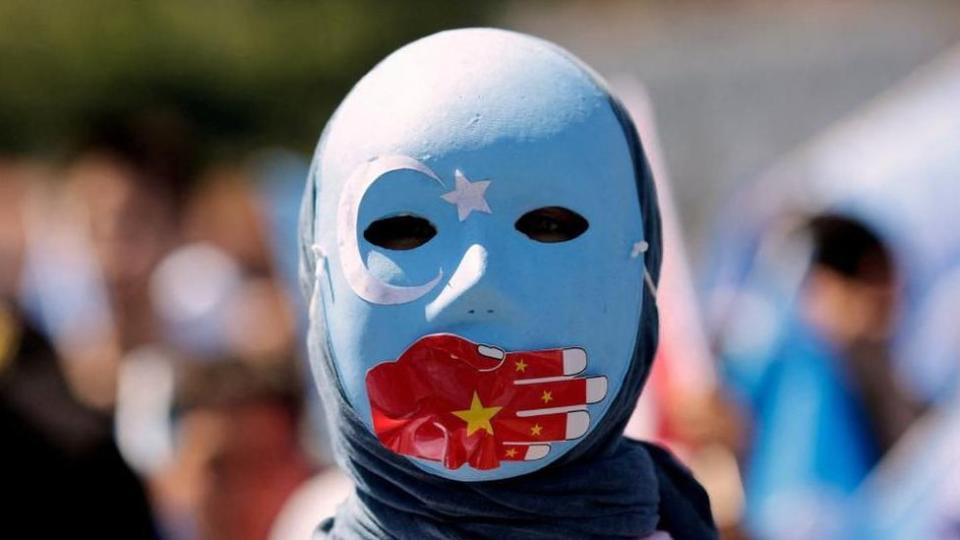 一名維吾爾族抗議者（他戴著飾有星月符號的藍色面具，一隻紅手舉著中國國旗捂住嘴）
