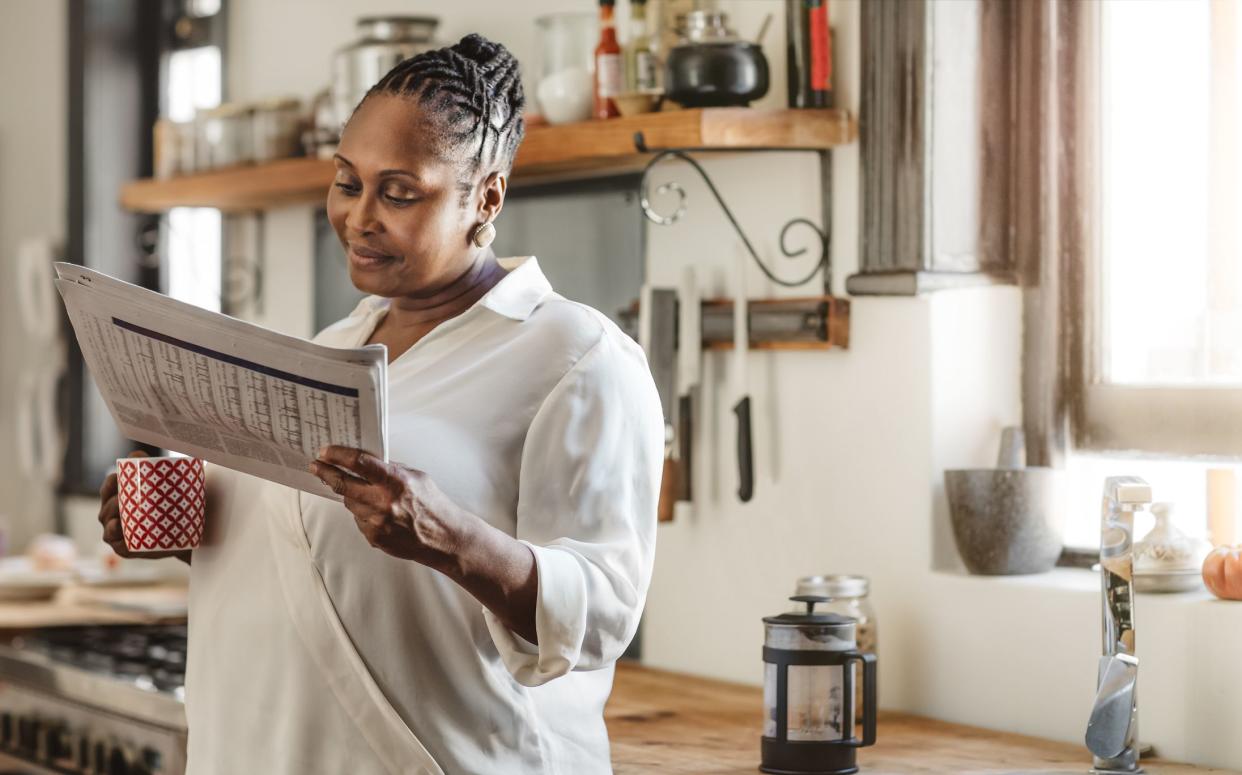 Smiling woman reading newspaper while also holding a coffee cup in her kitchen, very modern design with items on the counter