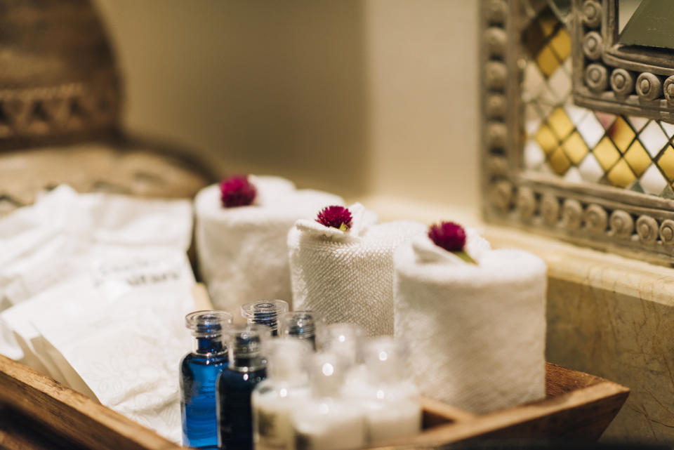 Rolled white towels with small flowers on top, and assorted spa products in small bottles arranged on a wooden tray. Mirrored decor in background