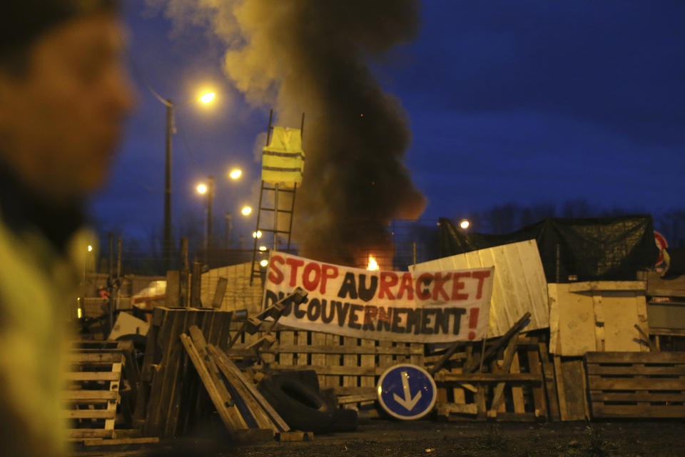 A demonstrator stands in front of a makeshift barricade set up by the so-called yellow jackets to block the entrance of a fuel depot in Le Mans, western France, Tuesday, Dec. 5, 2018, with banner reading "Stop the Government racket". French government's decision to suspend fuel tax and utility hikes Tuesday did little to appease protesters, who called the move a "first step" and vowed to fight on after large-scale rioting in Paris last weekend. (AP Photo/David Vincent)