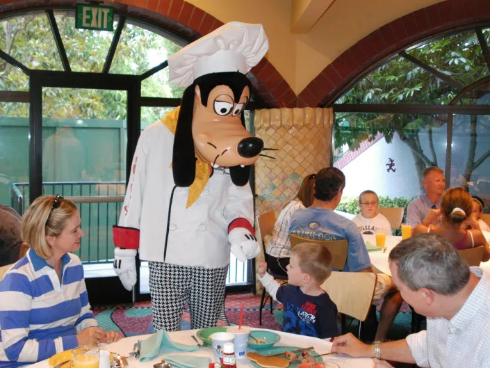 Someone in a Goofy dog costume in a chef's hat fist bumping a child sitting at a table in front of his breakfast with his family.