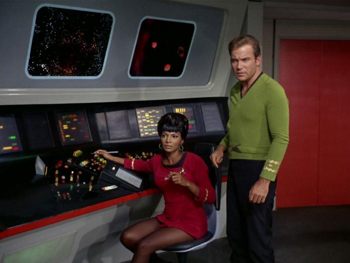 Nichelle in a red dress uniform and William in a green shirt universe. Both are at a control panel.