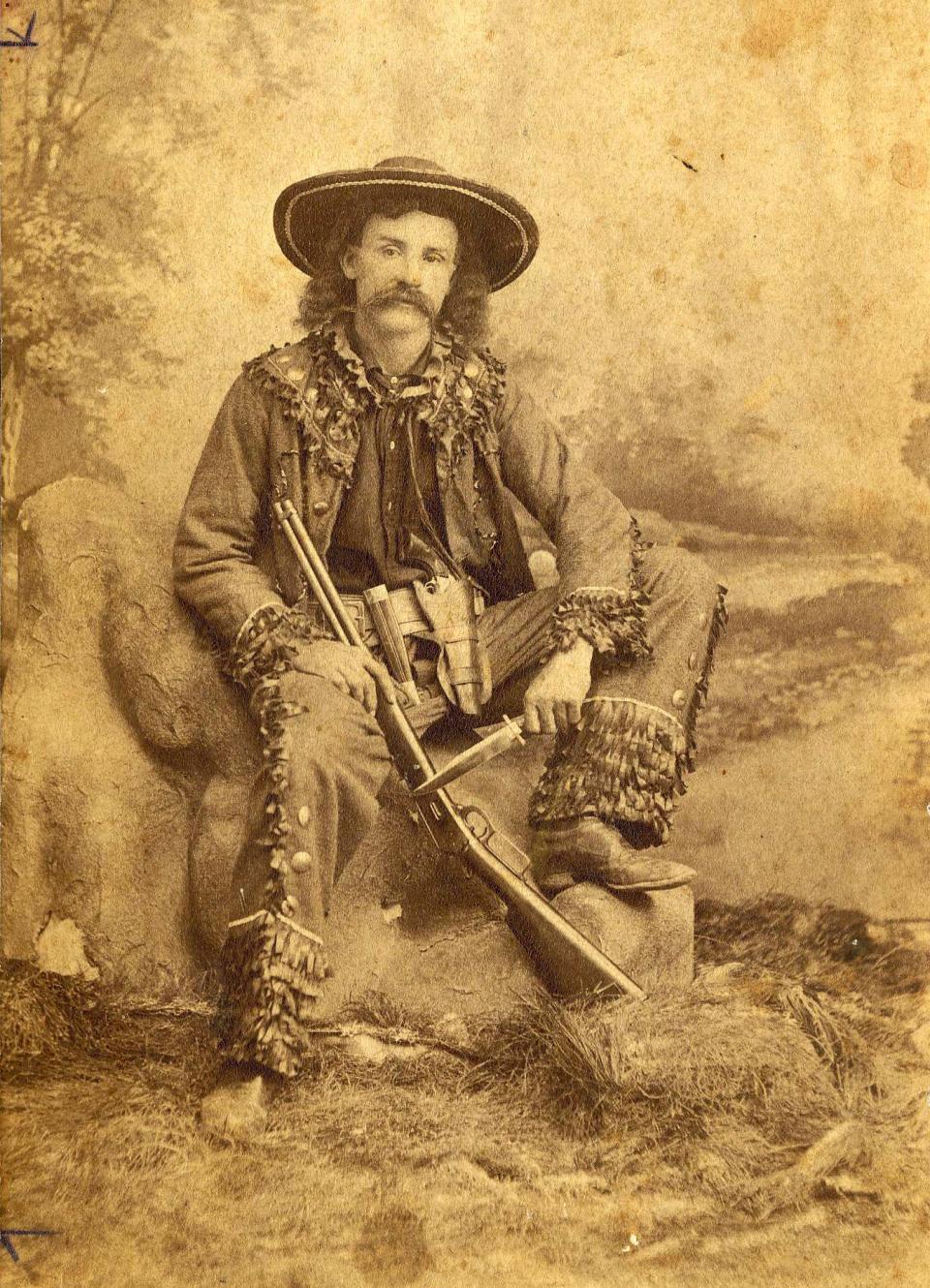 Circa 1880 photograph of Andrew Jackson Sowell, a Texas Ranger, holding a rifle by the barrel in his right hand, and a knife in his left. Portrait was taken in a studio with a painted background depicting a landscape. 