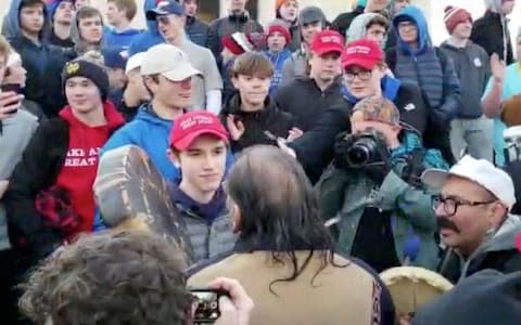 A teenager wearing a "Make America Great Again" hat stands in front of an elderly Native American singing and playing a drum in Washington - Credit: Survival Media Agency