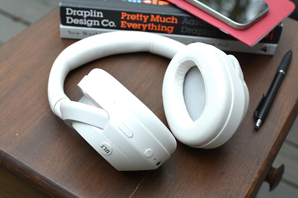 <p>White headphones sitting on a wooden table from above with a red notebook and phone in the background sitting on top of a black and a white book.</p>
