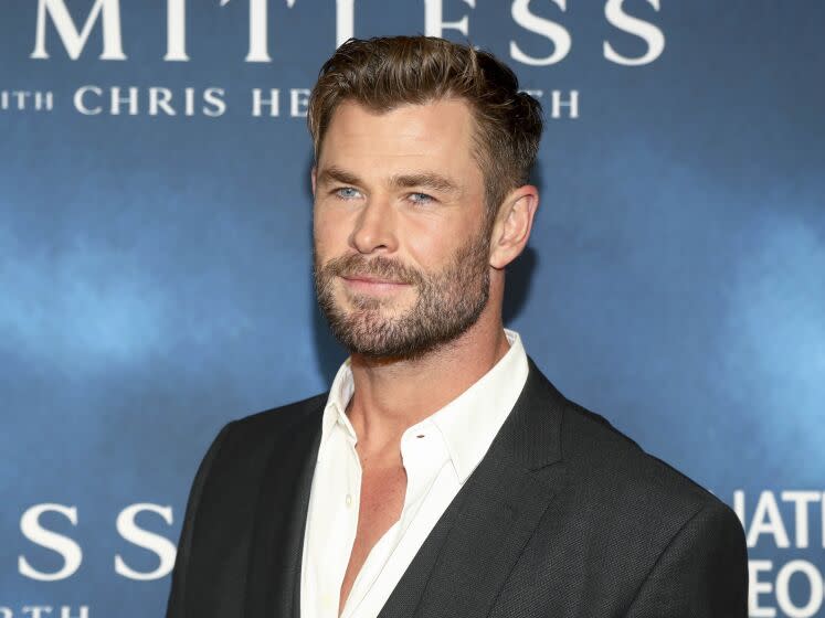 Chris Hemsworth in a black blazer and white suit shirt posing in front of a blue backdrop