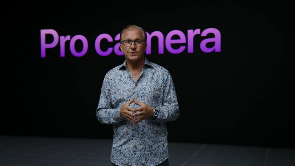 Fotograma del evento del iPhone de otoño de 2022. El vicepresidente de Apple, Greg Joswiak, se encuentra frente a un fondo oscuro con letras moradas que dicen: 