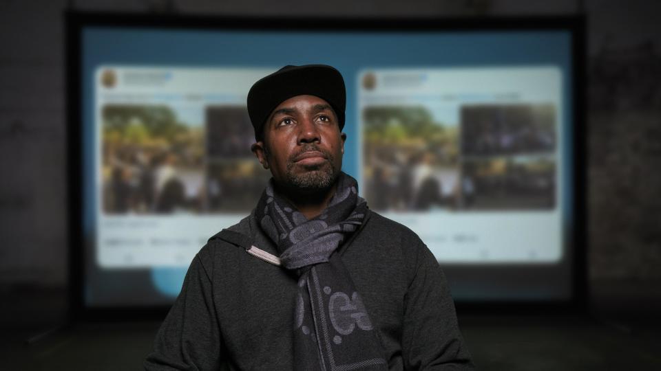 Director Prentice Penny, wearing a scarf and hat and with a screen projecting Tweets in the background.