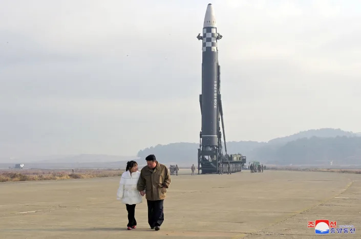 Kim Jong Un walks hand in hand across a flat expanse with a young woman in a white puffer jacket, a rocket with gray-and-white checkered midsection ready to launch behind them.