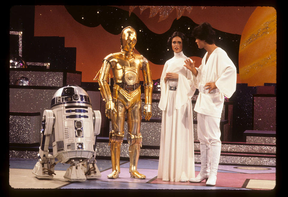 R2-D2, C-3PO, Carrie Fisher, and Mark Hamill are on a stage. Fisher and Hamill are dressed in white, iconic outfits from "Star Wars."