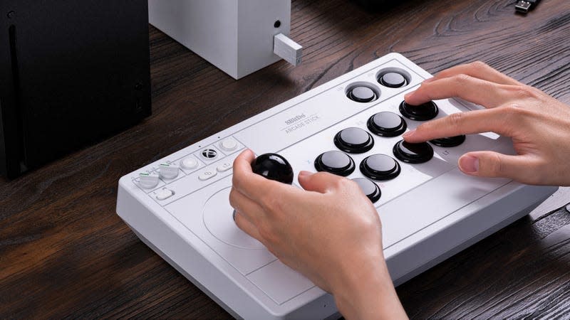 A user playing on the white version of the 8BitDo Wireless Arcade Stick for Xbox with multiple Xbox consoles pictured nearby.