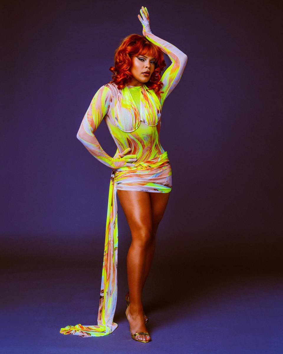 Vanessa Vanjie poses in a vibrant, patterned mini dress with long draped fabric, striking a confident stance on a plain backdrop