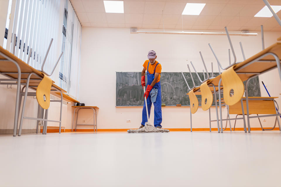 A janitor wearing overalls and a cap mops the floor in an empty classroom with upturned chairs on desks, focusing on cleanliness in a work environment