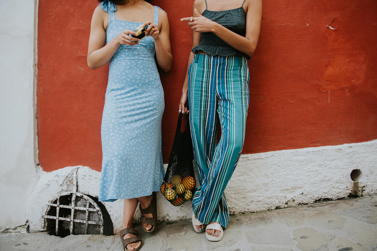 Conceptual and bright image of two females standing against a red wall in the streets of Positano. They are wearing fashionable and light clothing, appropriate for warm weather and comfortable walking sandals, perfect for exploring the winding streets of Italy.