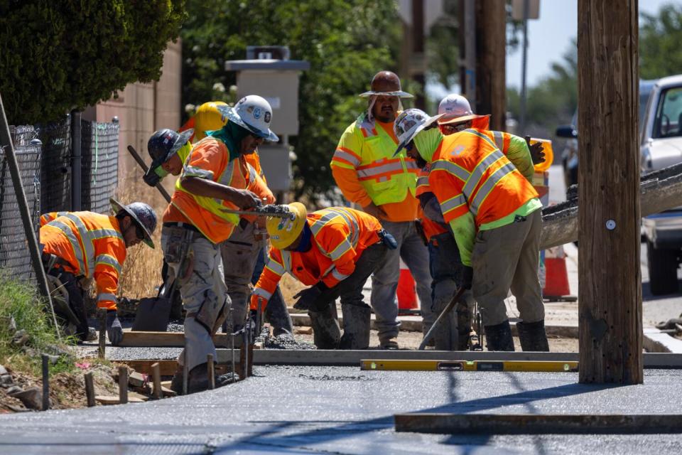 Construction workers in orange vests work on a sidewalk.