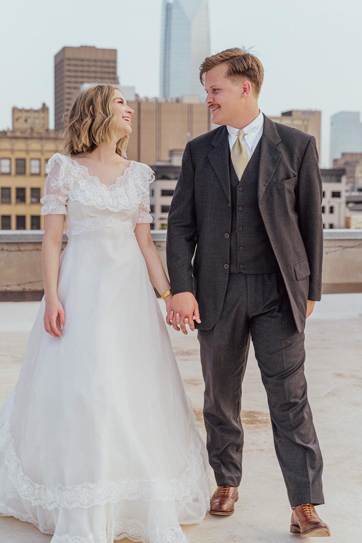 A bride and groom hold hands on top of a roof in their wedding attire.