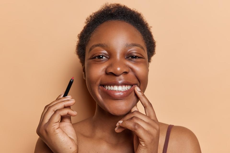 Happy dark-skinned woman with short hair holding a lip liner pencil in a peach-colored background