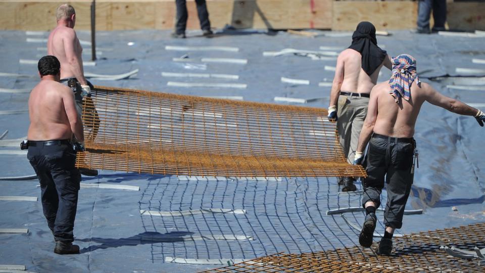 Arbeiter mit freiem Oberkörper tragen auf einer Baustelle ein Stahlgitter. Foto: Ole Spata