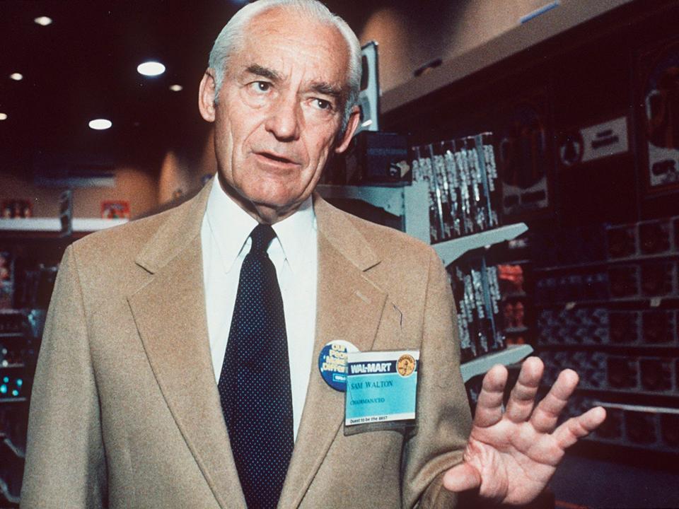 Sam Walton gestures and looks off camera while wearing Walmart nametag