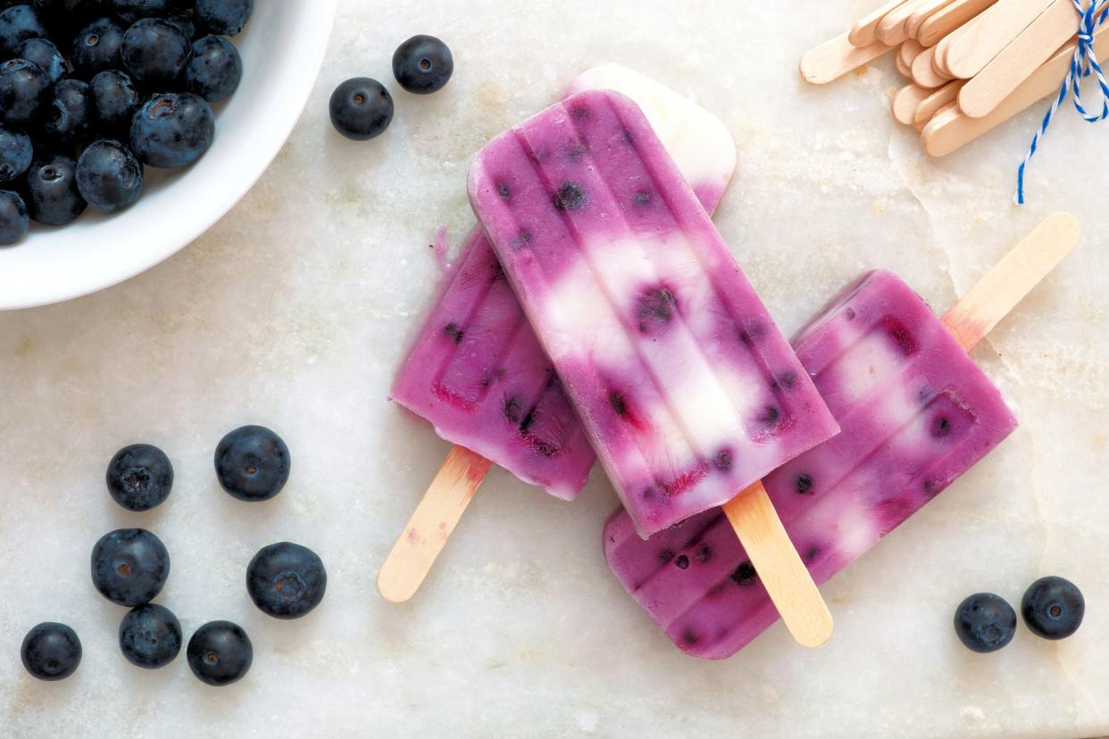 Three blueberry vanilla popsicles, one on top of two, surrounded by blueberries, some blueberries in the a white bowl on the top left, many popsicle sticks on the top right, on a white marble table
