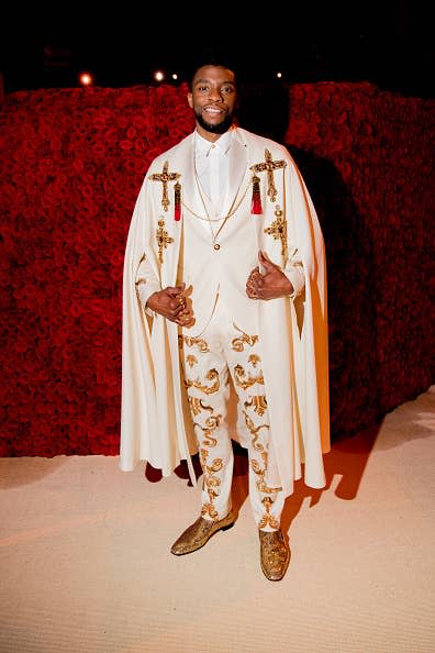 Chadwick Boseman in a white suit with gold patterns and a cape at a gala event
