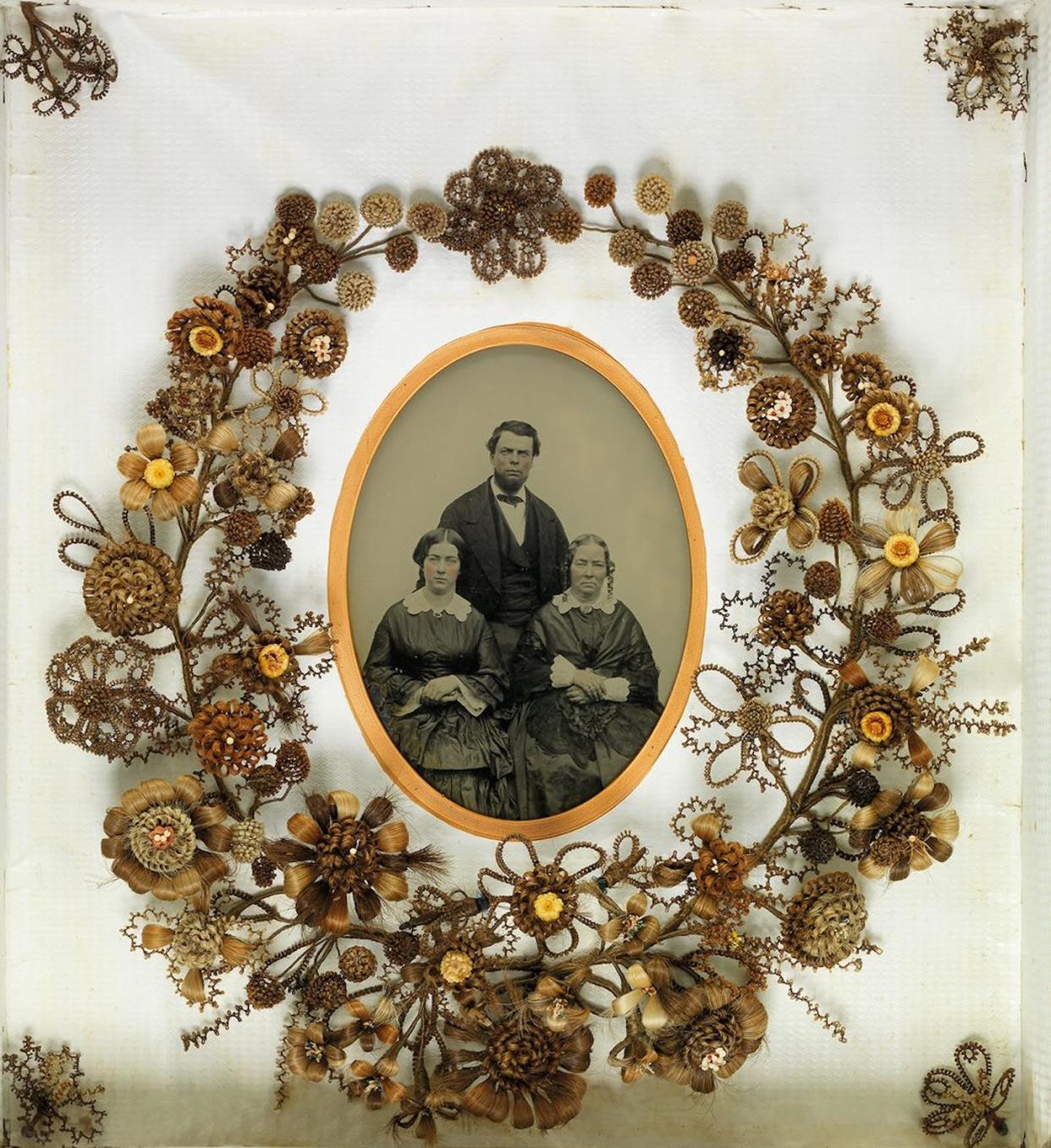 Mechones de pelo enrollados en forma de flor rodeando una foto en blanco y negro de una familia. Ambrotipo (de tres personas) y corona de flores. Alrededor de 1850. <a href="https://muttermuseum.org/exhibitions/woven-strands" rel="nofollow noopener" target="_blank" data-ylk="slk:Mütter Museum;elm:context_link;itc:0;sec:content-canvas" class="link ">Mütter Museum</a>