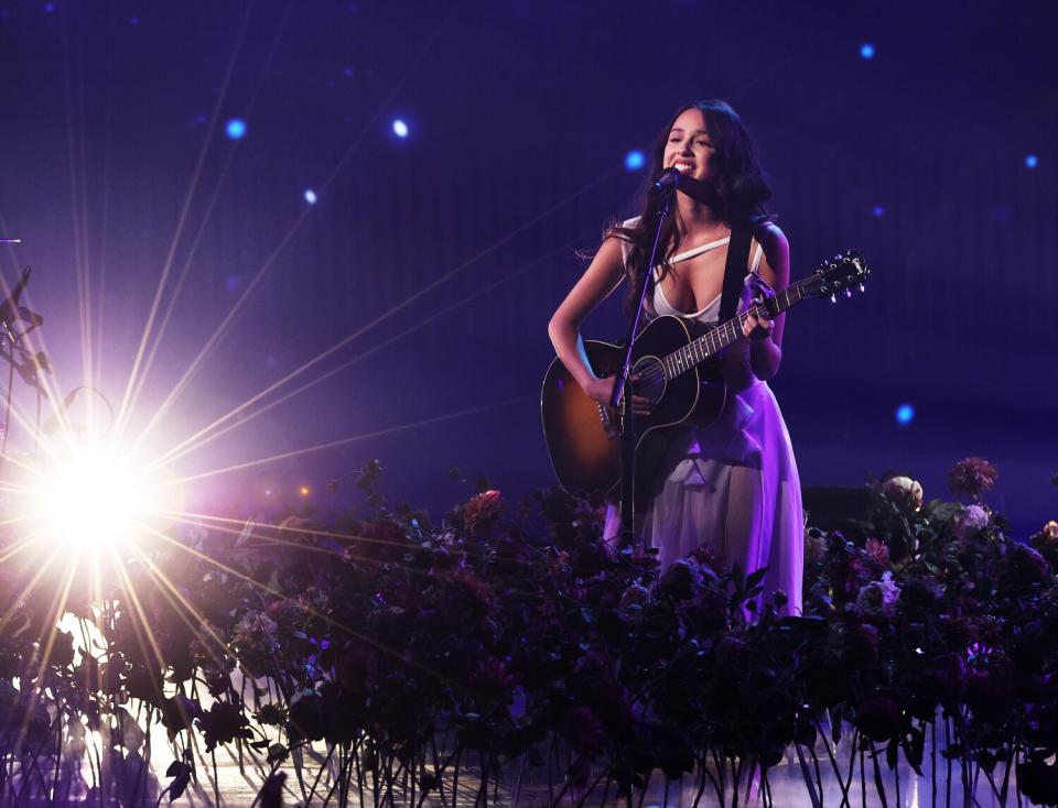 A woman in a purple dress sings and plays the guitar onstage.