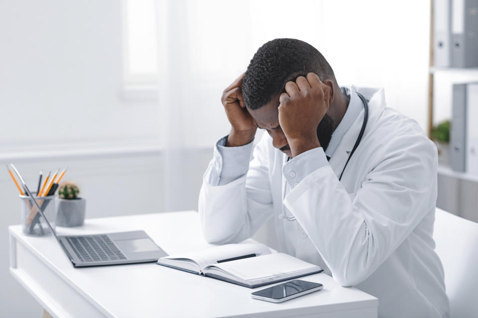 Exhausted young african doctor sitting in office in front of laptop, having headache, copy space