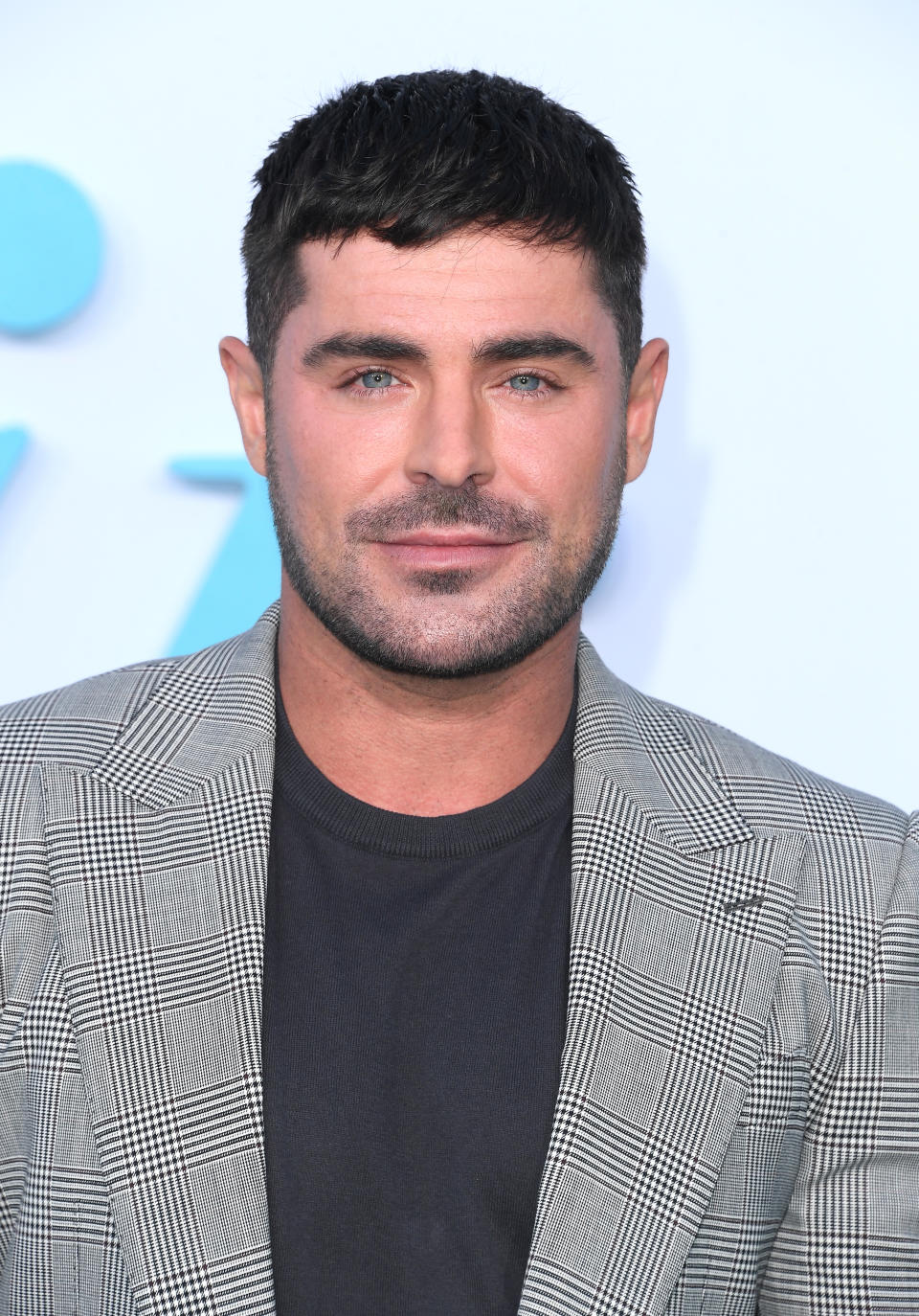 Zac Efron smiles at an event, wearing a checkered blazer over a black shirt