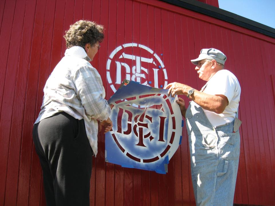 Richard Porath of Waltz and his wife, Denise, are shown using a large stencil to paint the letters "DT&I" in a circle on the side of the caboose. Porath, with help from others, recently completed most of the remaining work on the Flat Rock Historical Society's 99-year-old caboose. The public can tour the caboose today.