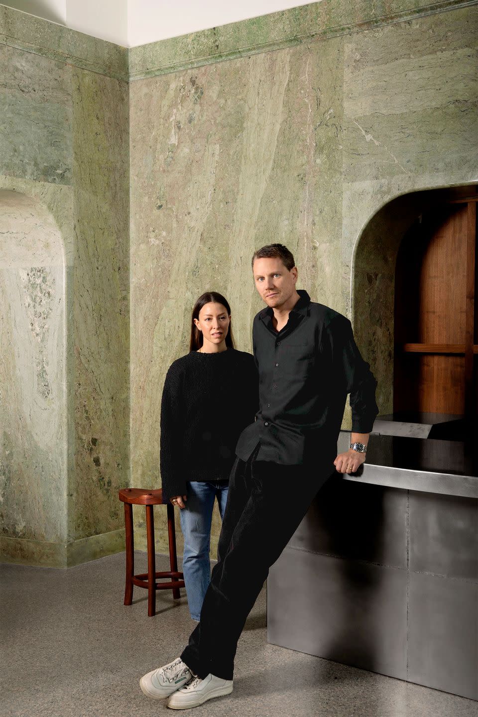 two people standing next to a metal counter and small wooden stool and marble clad walls