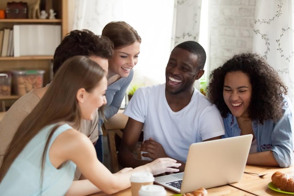 A group of friends smile and talk while looking at something on a laptop.