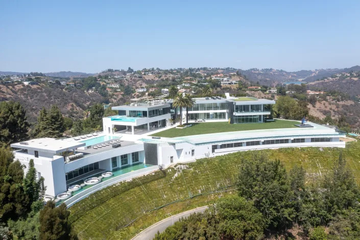 an aerial view of a white mansion, The One Bel Air, and its pools, surrounded by hills and green grass