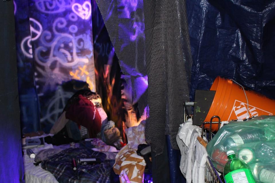 The first room in the art installation, which has a tent, a grocery cart filled with recyclables and a pile of backpacks leads into a small graffitied room with a mattress and piles of clothes. 