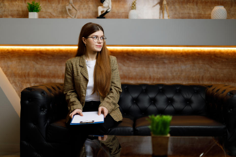 A woman with long hair sits on a black leather couch, holding a clipboard. She wears glasses, a plaid blazer, and a white shirt. She looks to her left