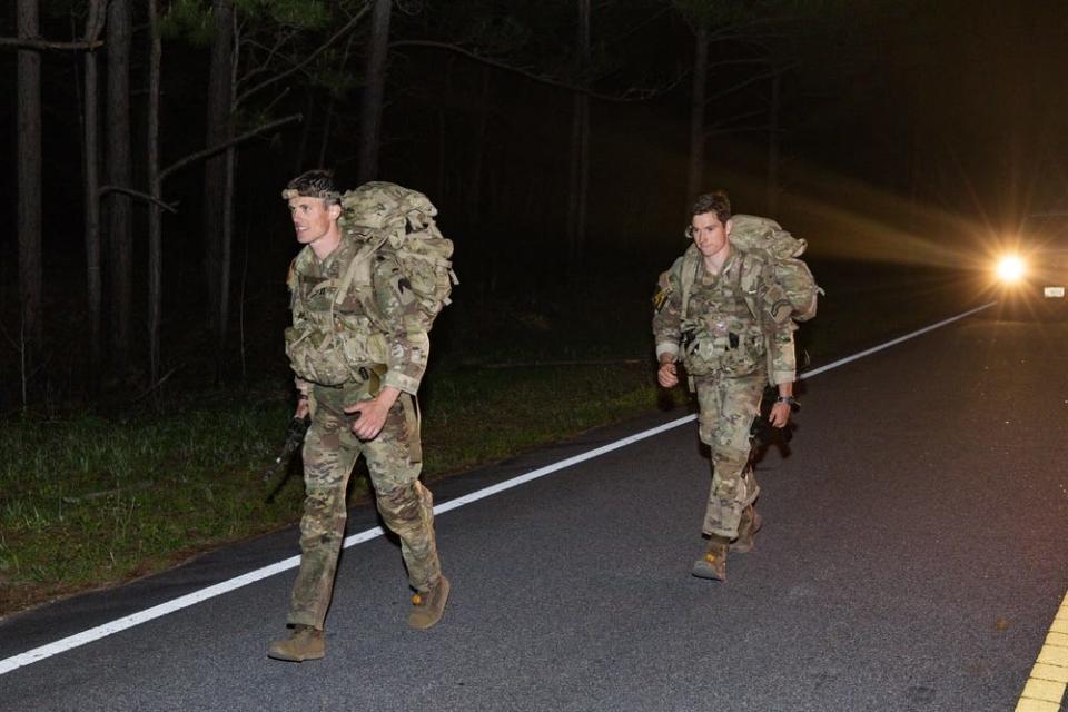 US Army soldiers complete a ruck march at night illuminated by the headlights of a vehicle behind them