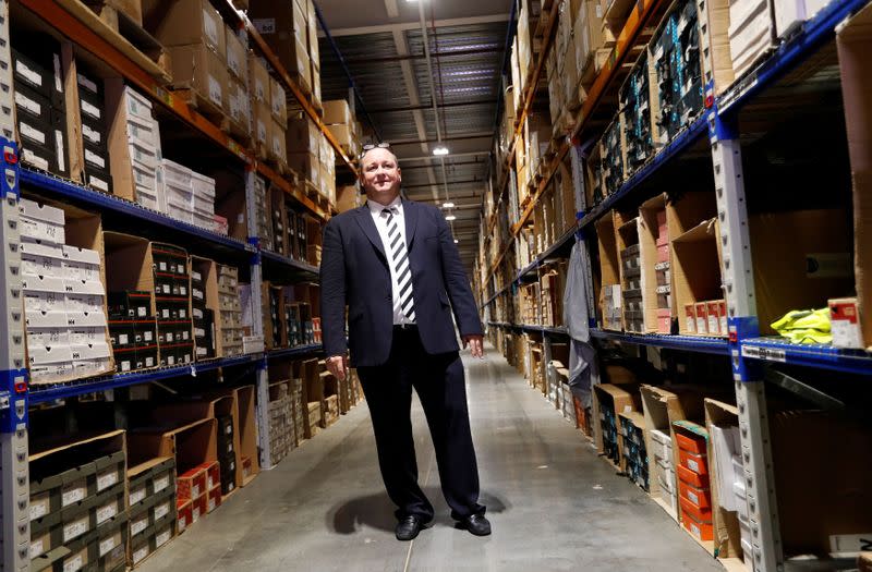 FILE PHOTO: Mike Ashley, founder and majority shareholder of sportwear retailer Sports Direct, stands between shelves of stock during a factory tour after the company's AGM, at the company's headquarters in Shirebrook