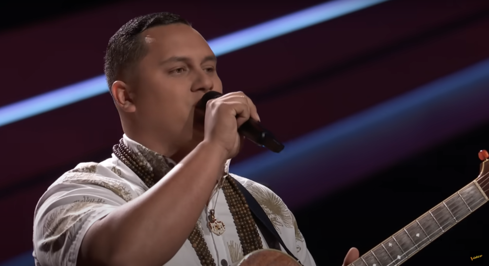 Kamalei  with a microphone singing and playing an acoustic guitar on stage