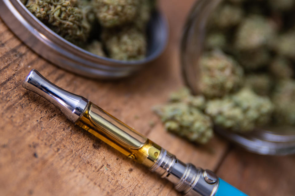 THC oil concentrate filled vape pen on natural wood with an open glass container full of Mango Kush strain marijuana buds grown &amp; sold in dispensaries through-out Southern California.