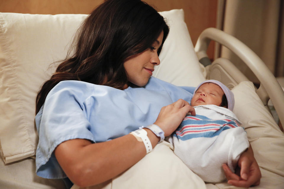 Gina Rodriguez lies in a hospital bed smiling at a newborn baby wrapped in a blanket, cradled in her arm