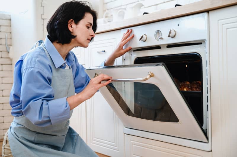 Woman opening door of electric oven, looking inside and check readiness of dish.