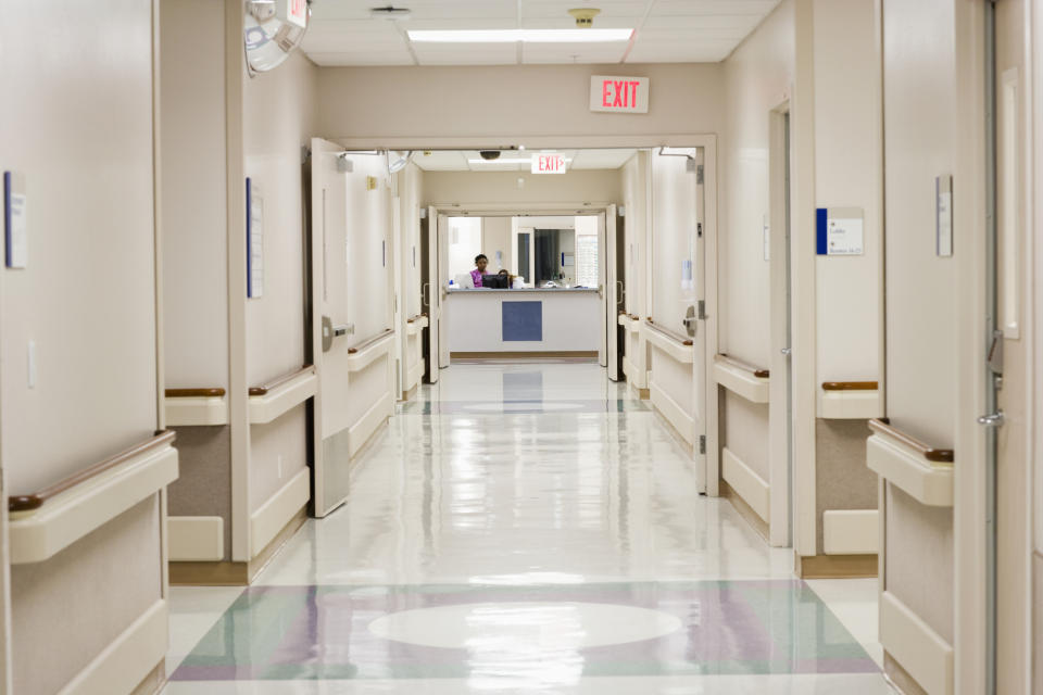 A quiet hospital hallway with shiny floors, handrails, and an exit sign in the background. Two healthcare workers are seen at the nurse's station at the end of the hall